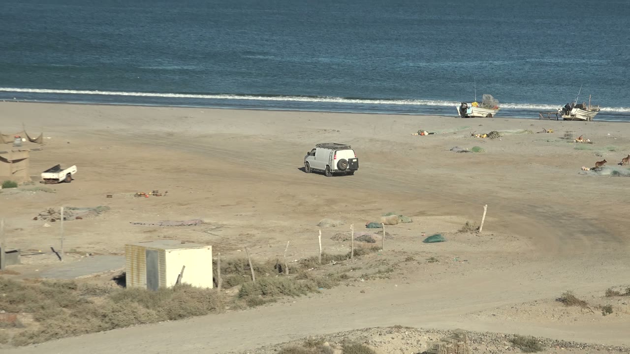 Opening wide view of San Juanico beach in Baja California Sur with a sandy shore, small boats near the water, a white van on the beach, a dirt road, scrub vegetation, distant landforms, and no visible people.