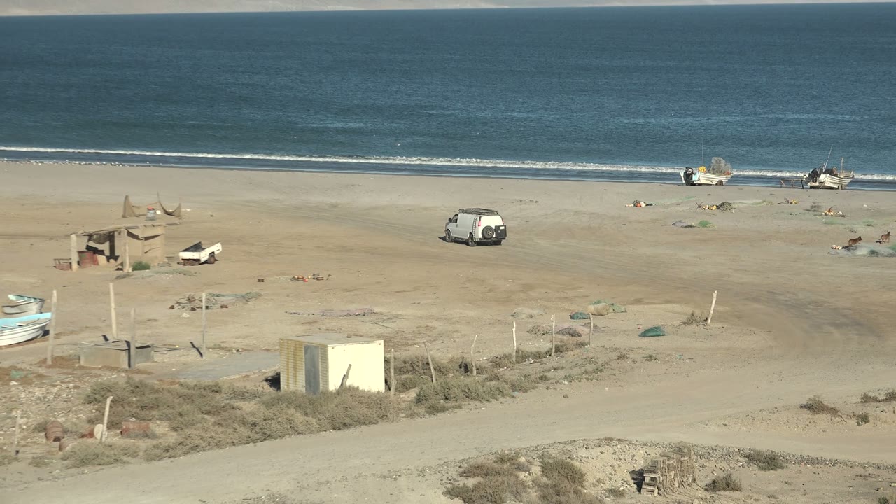 Sandy beach and blue ocean with beached boats, a van, and small structures.