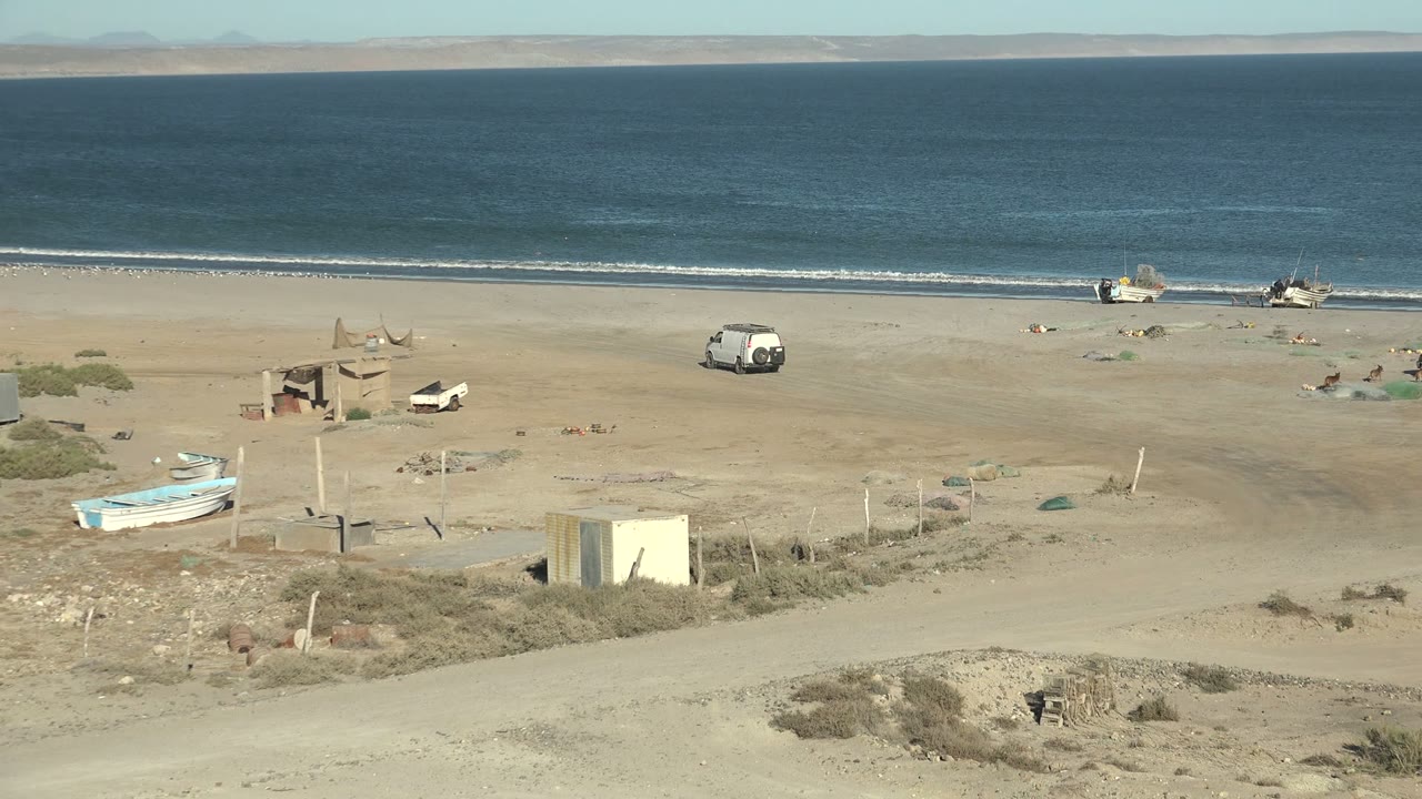 San Juanico coastline view with the ocean horizon high in frame, boats clustered near the waterline, a van and other small vehicles on the sand, a foreground dirt road, dry vegetation, and no visible people.