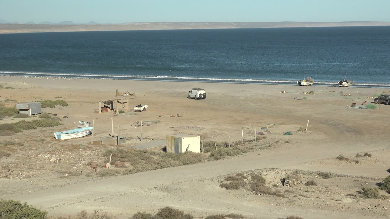 Beach panorama with boats by the shore, parked vehicles, and scrub foreground.