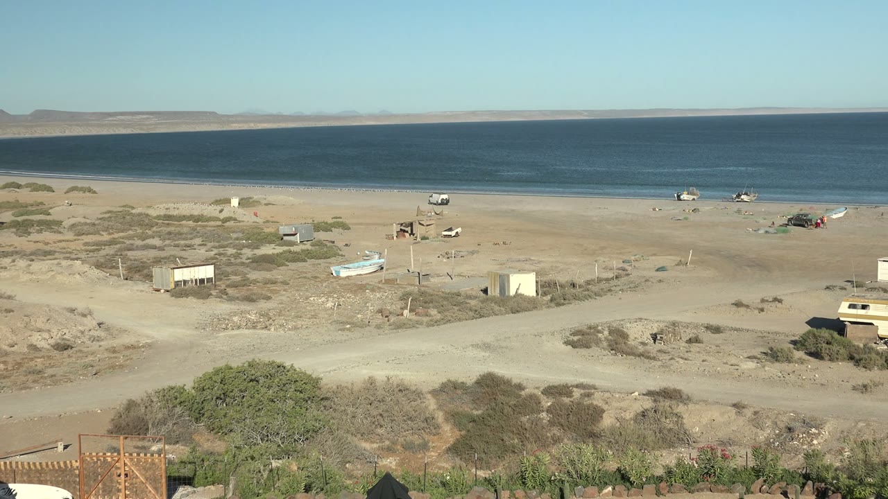 Wide view of San Juanico, Baja California Sur showing a sandy desert beach, calm blue ocean, small boats near shore, scrub vegetation, dirt tracks, partial foreground camp structures, and no visible people.