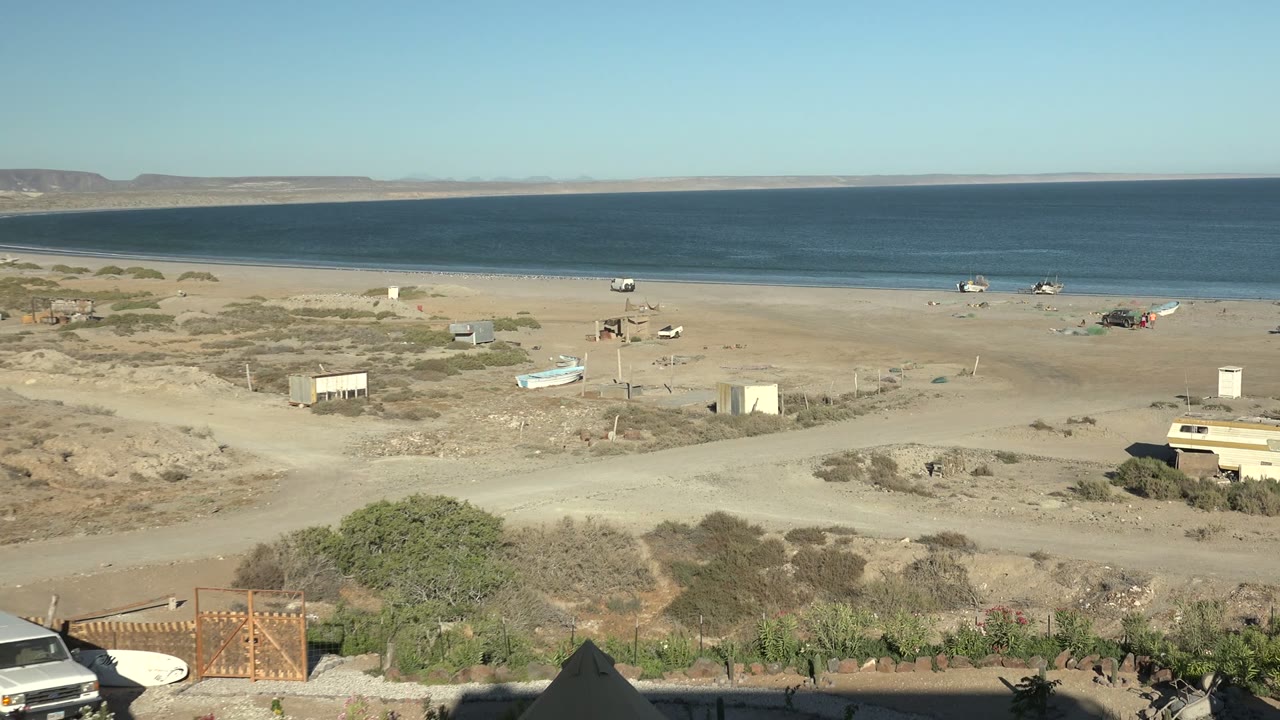 Sandy beach and ocean with boats, scrub, dirt road, and a vehicle beginning to appear below.