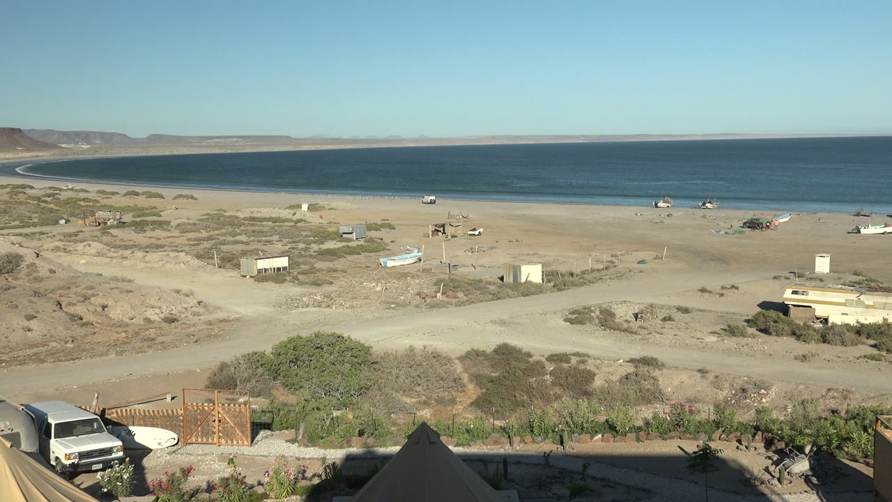 Wide San Juanico coastal view with a white vehicle more visible at the bottom edge, wooden gate and wall in the foreground, sandy beach, small boats, calm ocean, scrub patches, and no visible people.