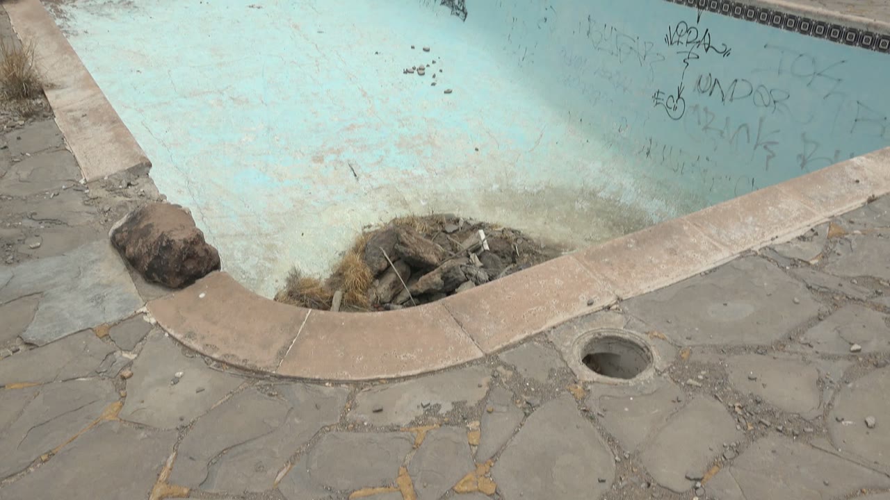Early frame of an abandoned pale blue swimming pool in Mulege, Baja California Sur, with graffiti on the wall, debris near the deep end, cracked stone decking, and no visible people.