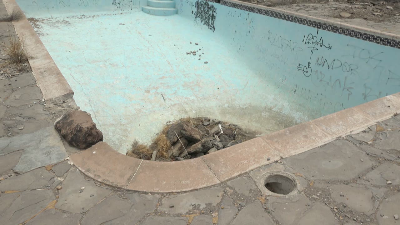 Graffiti-marked empty pool with debris and dry weeds at the bottom edge.