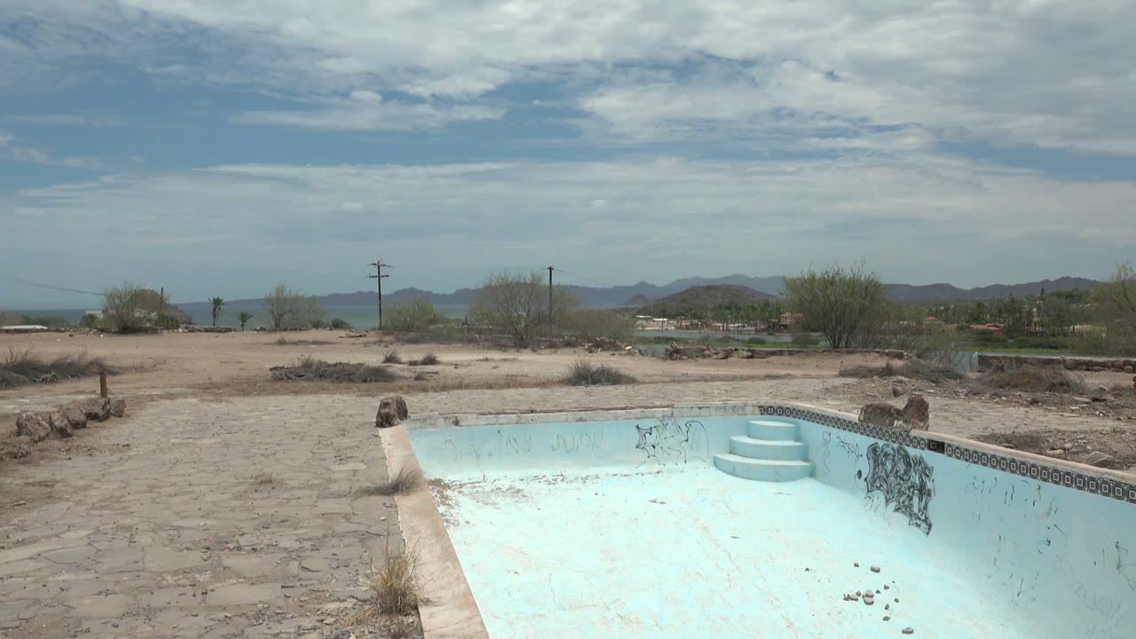 An empty pale blue swimming pool sits in a dry yard in Mulege, Baja California Sur, with sandy ground, sparse shrubs, distant mountains, and no visible people.