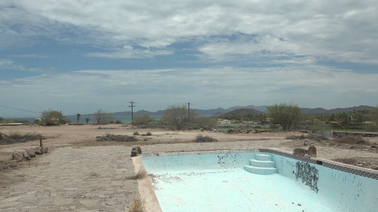A wider view of the empty pool in Mulege, Baja California Sur shows the blue basin, rough pool edge, sandy open ground, utility poles, distant mountains, and no visible people.