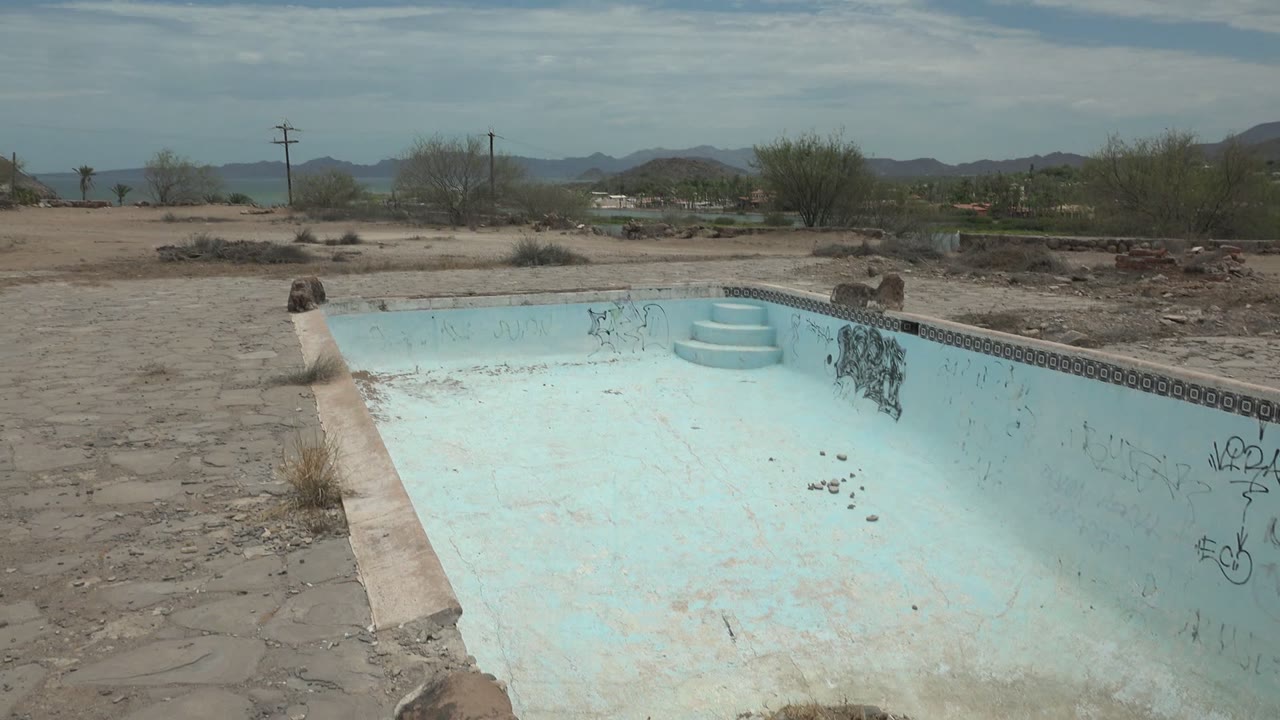 Opening frame of an abandoned pale blue pool in Mulege, Baja California Sur, with graffiti on the far wall, stone paving, desert shrubs, distant hills, and no visible people.