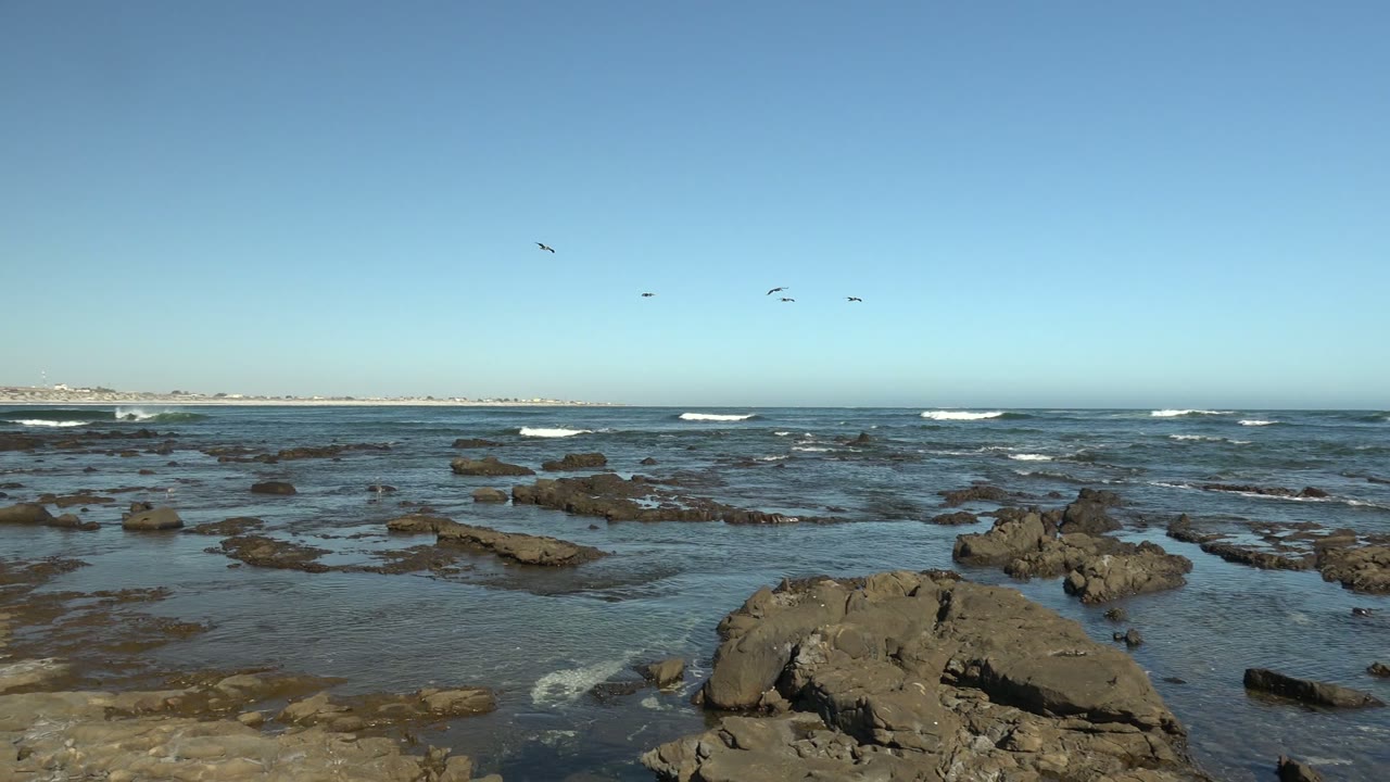 Rocky La Bocana coast with scattered tide pools, a low ocean horizon, small breaking waves, distant birds at the right side of the sky, and no visible people.