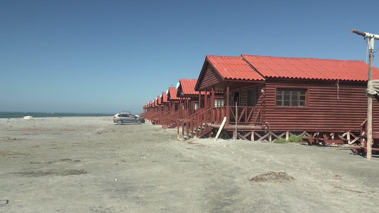 At 0:00 in La Bocana, red-roofed wooden cabins line the sandy beach with ocean at left, a gray car in the distance, and no visible people.