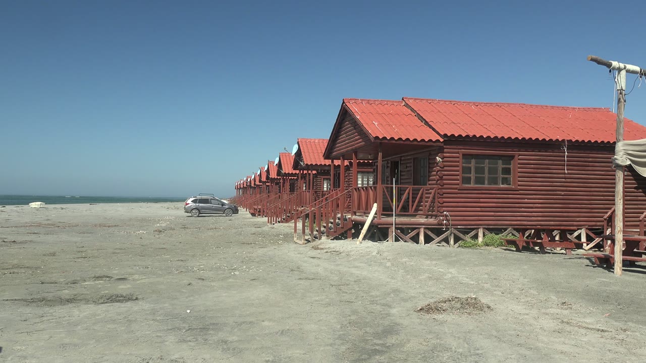 At 0:02 in La Bocana, the nearest cabin fills more of the right side, the ocean remains visible on the left, and no visible people appear on the sand.