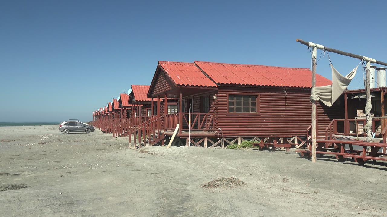 Opening frame of red wooden beach huts on sand in La Bocana, Baja California Sur, with a white car at the left near the ocean and no visible people.