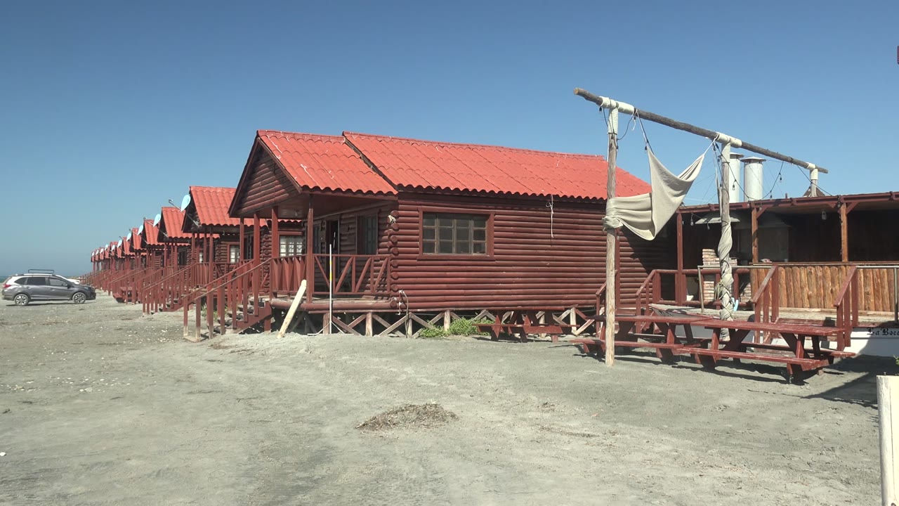 Red wooden cabins in La Bocana, Baja California Sur line the sandy ground as a dark car appears near the left edge, with no visible people.