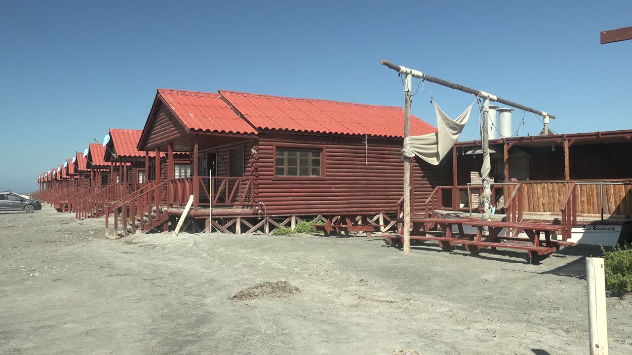 Red wooden cabins and porch structures fill the midground in La Bocana while the car is nearly out of view on the left, with no visible people.