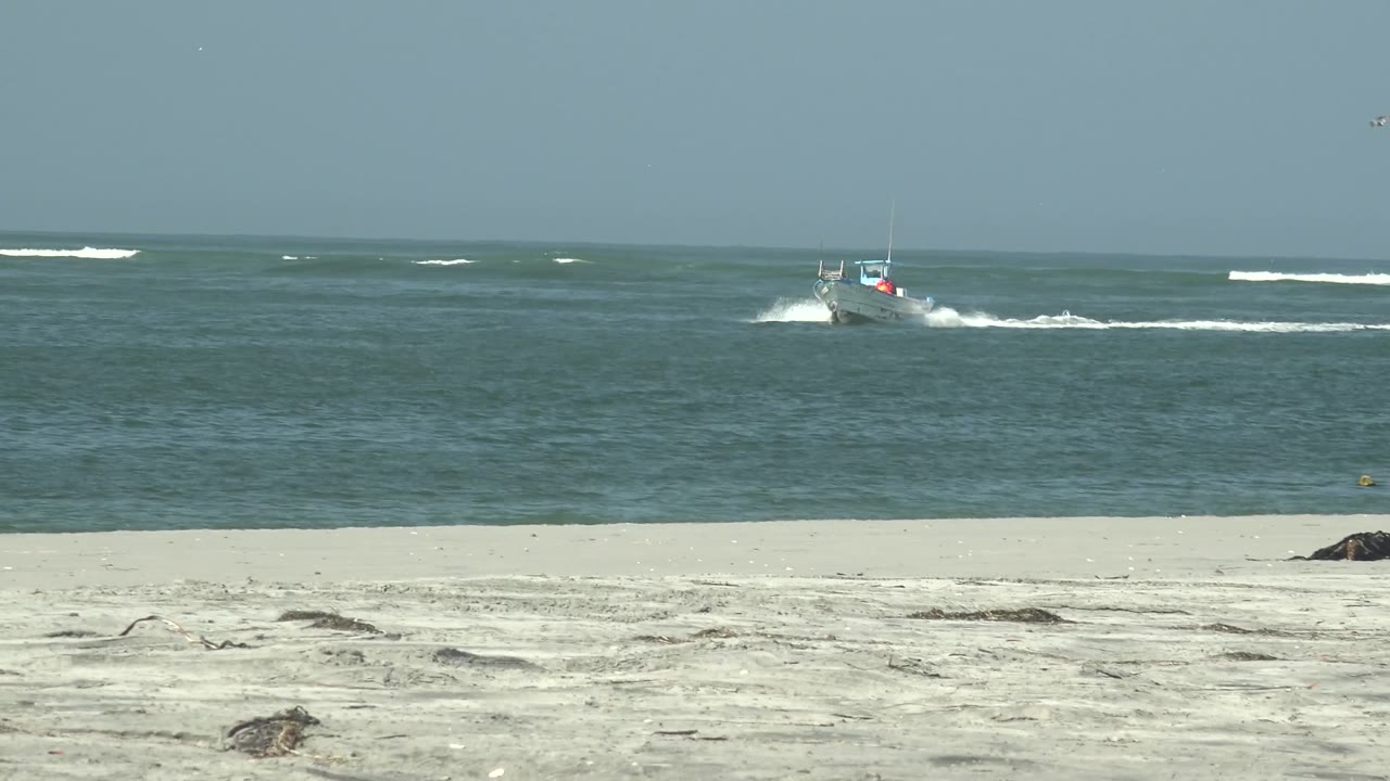 A small boat moves on blue-green water off a sandy beach in La Bocana, Baja California Sur, with white surf behind it and no visible people.