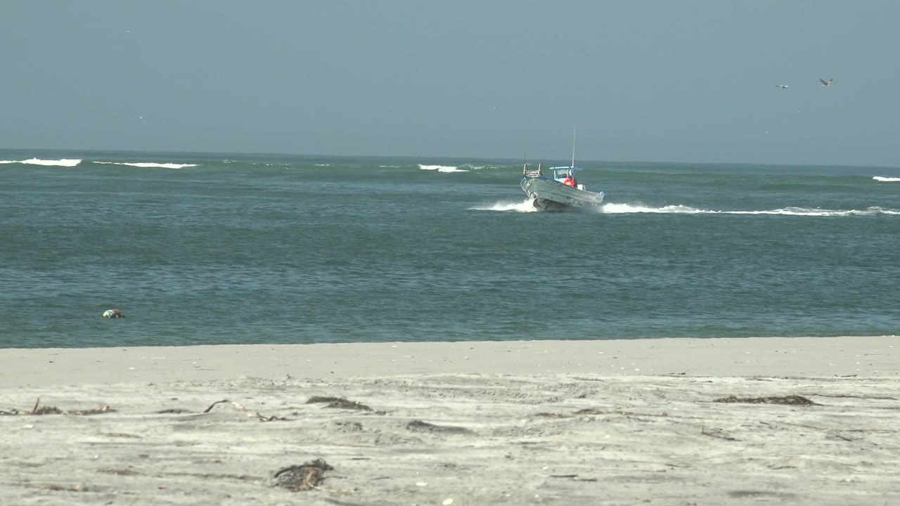 A small boat runs near the right side of the La Bocana ocean frame, leaving white spray across blue-green water above the sandy beach, with no visible people.