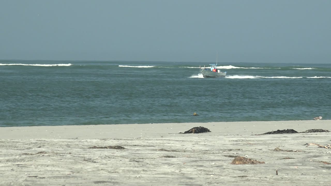 A small boat appears farther right offshore at La Bocana, Baja California Sur, cutting through blue water with a bright wake, while pale sand and low white surf fill the frame and no people are visible.