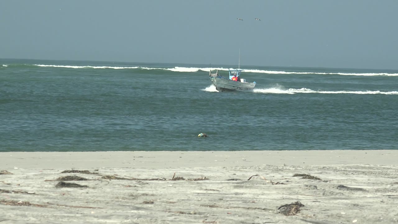 At La Bocana, Baja California Sur, a small boat runs across the midground water beyond pale sand and scattered driftwood, with white surf behind it and no visible people.