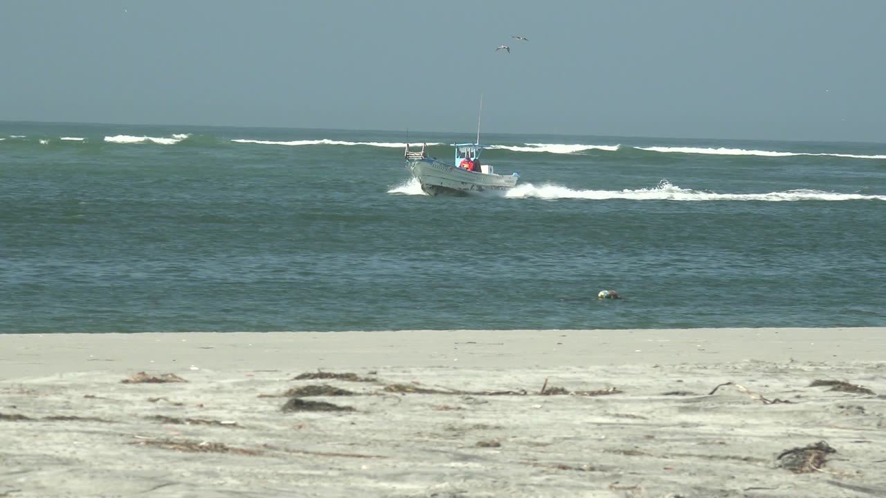 At La Bocana, Baja California Sur, the small boat sits farther to the right of frame with bright wake on darker water, pale beach below, broken surf behind, and no visible people.