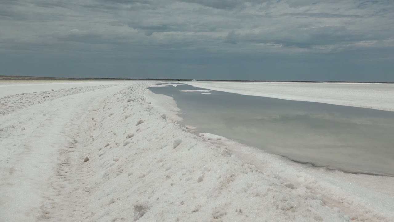 Snow-covered path with footprints beside partially frozen water in Guerrero Negro, Baja California Sur, with a low horizon and no visible people.