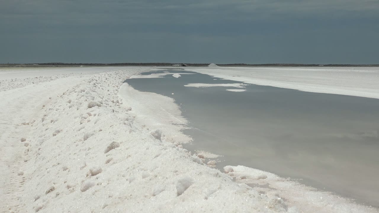 Opening frame in Guerrero Negro, Baja California Sur showing a rough white path with footprints beside a gray frozen water surface, flat horizon, muted sky, and no visible people.