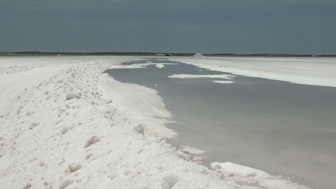 Frame at 0:02 in Guerrero Negro, Baja California Sur showing the white path closer to center, gray frozen water to the right, a low dark horizon, and no visible people.