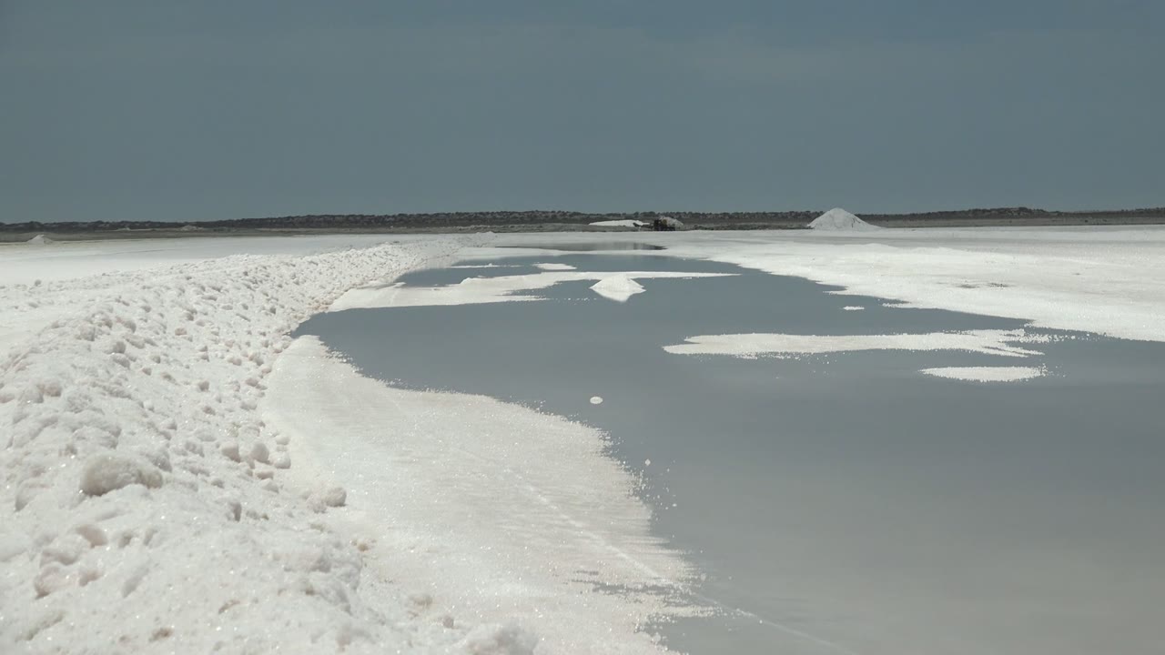 White salt crust surrounds shallow gray water pools on a Guerrero Negro salt flat, with low distant salt piles and no visible people.