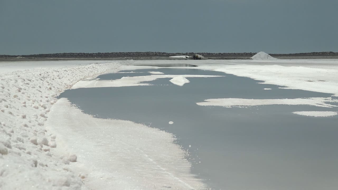 Irregular water pools break up the white Guerrero Negro salt flat, with a white salt pile near the distant horizon and no visible people.