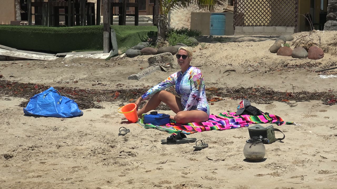 At 0:00 in San Carlos, Baja California Sur, a woman in sunglasses sits on a striped towel on the sand with an orange bucket, blue bag, shoes, and a container around her.