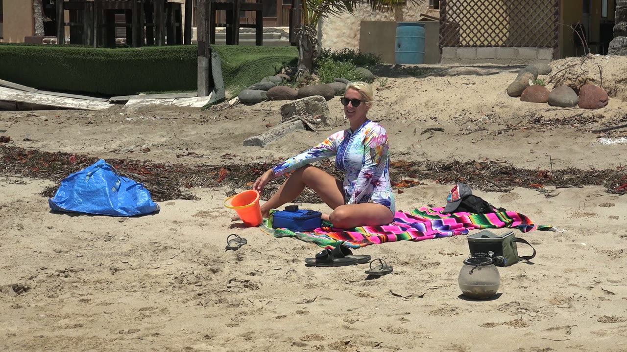 Seated woman reaches toward an orange bucket on the sand beside a striped towel.