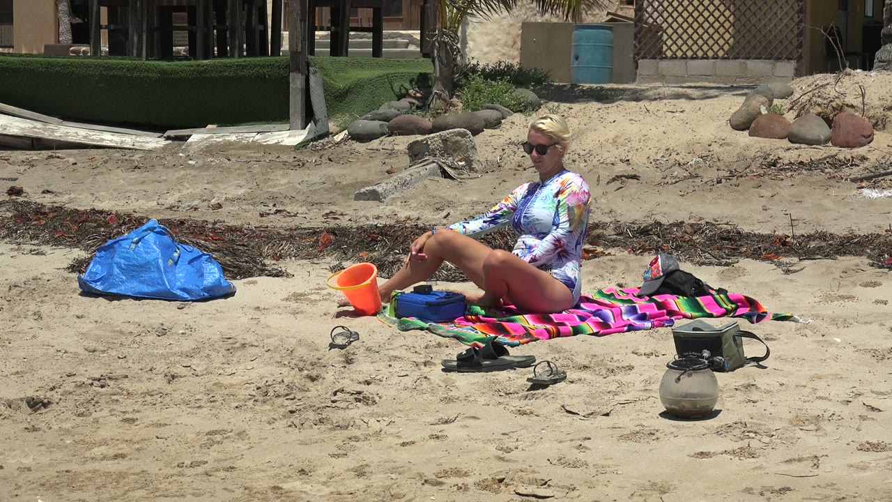 At 0:02 in San Carlos, Baja California Sur, the woman remains seated beside the orange bucket, with the blue bag left of the towel and a green fence and structures behind the sandy area.