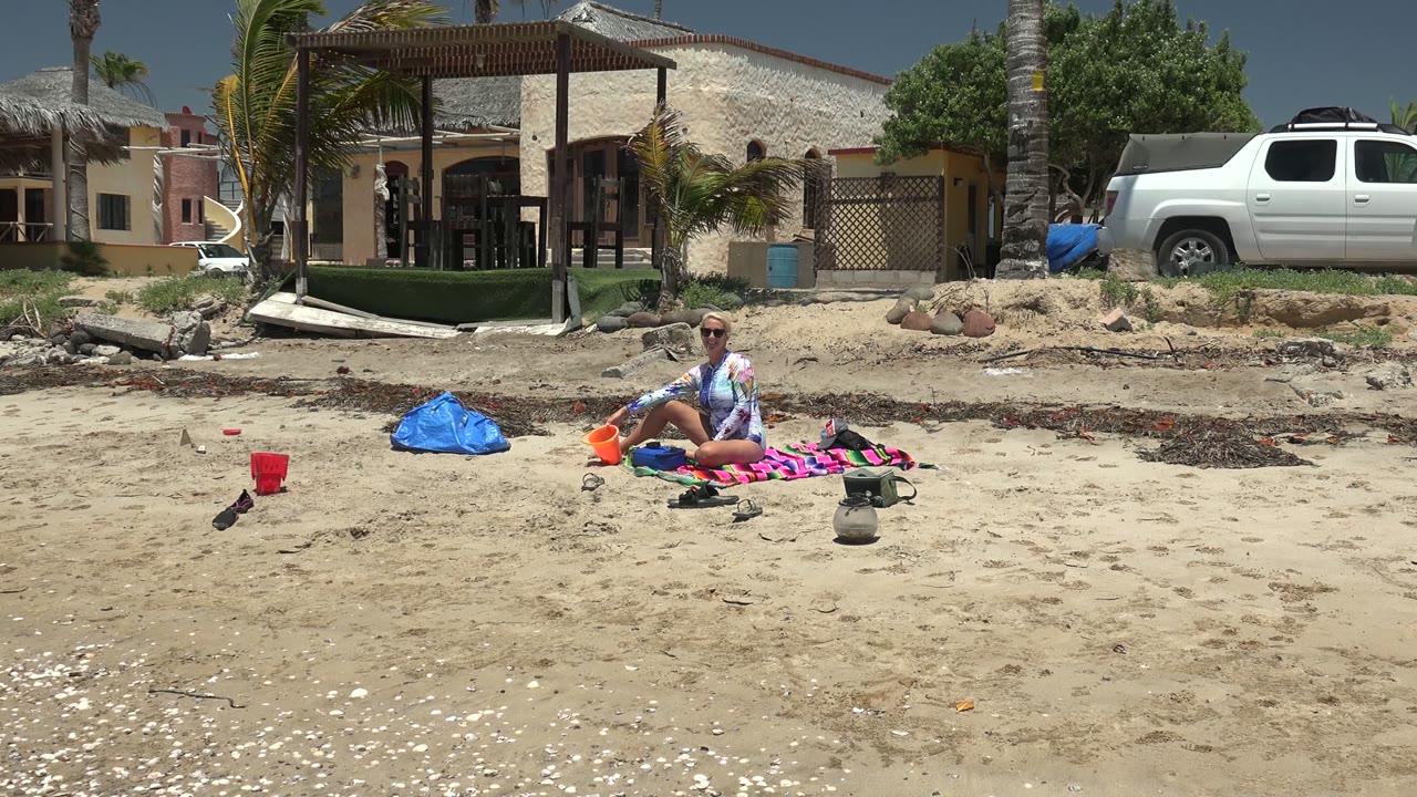 At 0:00 in Puerto San Carlos, a person sits on a striped towel beside a blue bag and red cup, with palm trees, a thatched-roof building, and a white truck behind the sandy beach.