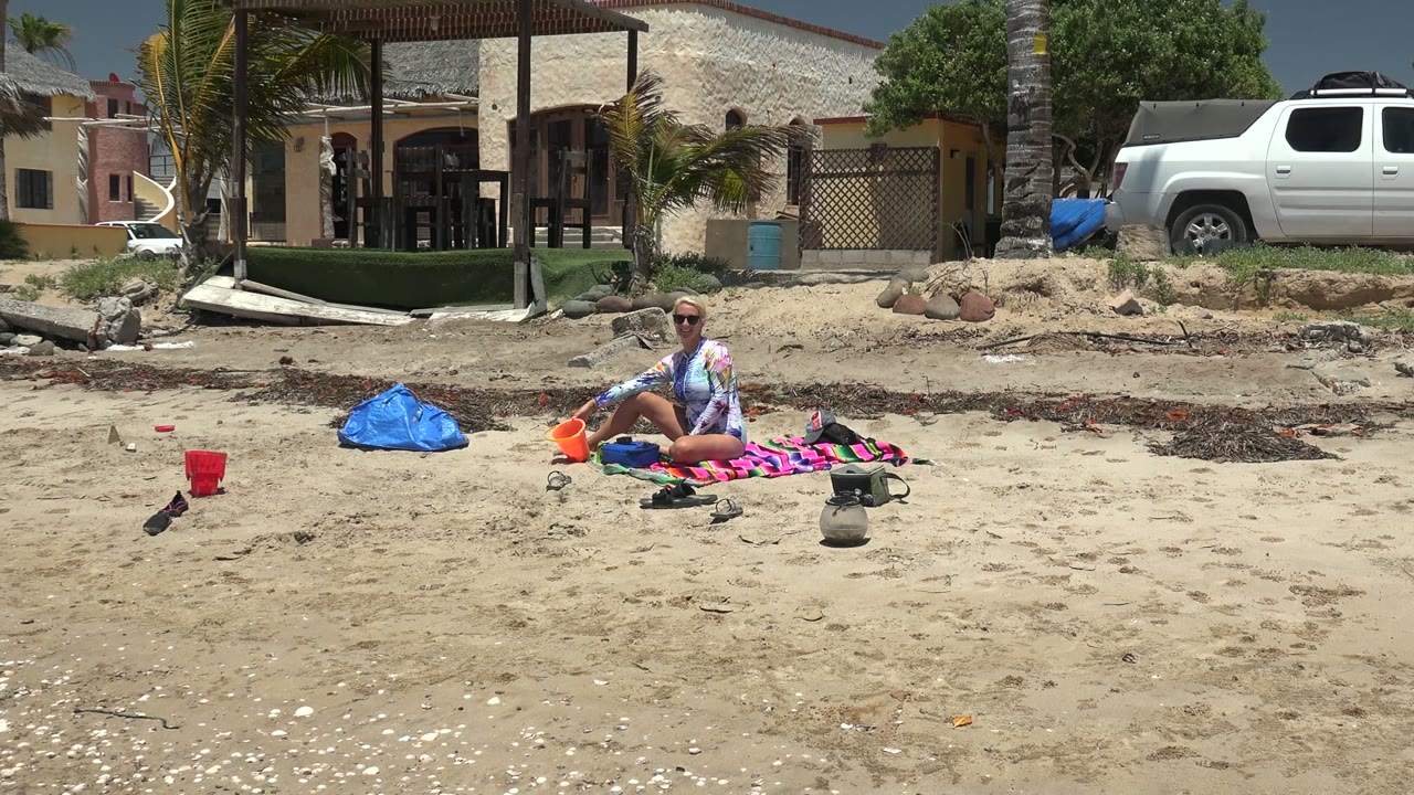 Beachgoer on a colorful towel with an orange bucket and blue bag on sandy shore.