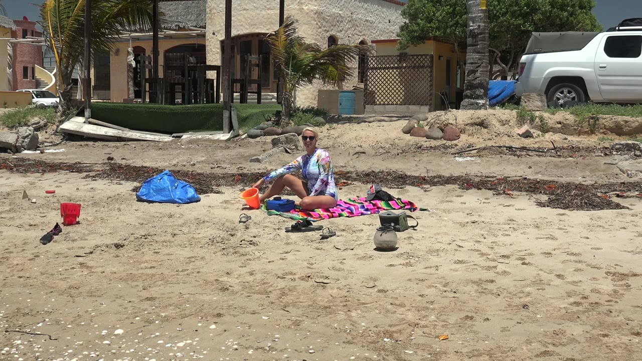 At 0:02 in Puerto San Carlos, the beachgoer sits upright on the striped towel, with a blue bag to one side, a red cup in the sand, and the white truck and building spanning the background.