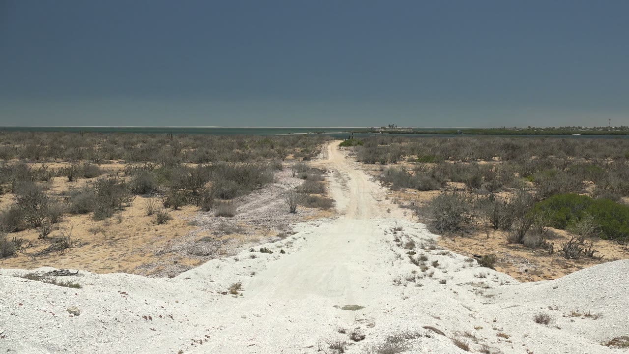 The road continues through low arid vegetation in San Carlos, Baja California Sur, Mexico, with a flat horizon and no visible people.