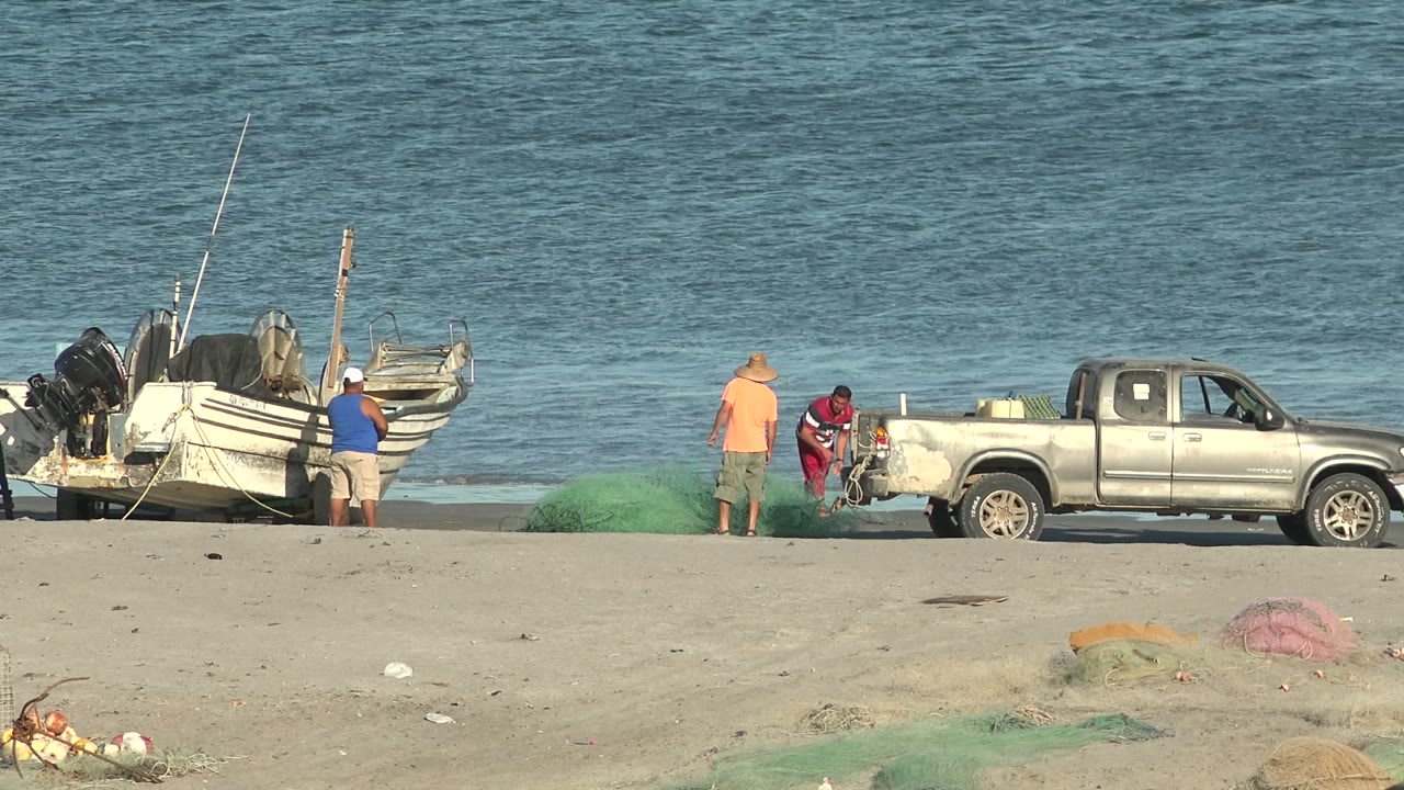 At the start of the San Juanico clip, three men stand near green fishing nets, a silver pickup truck, and a small boat on the sandy shore with blue ocean behind them.