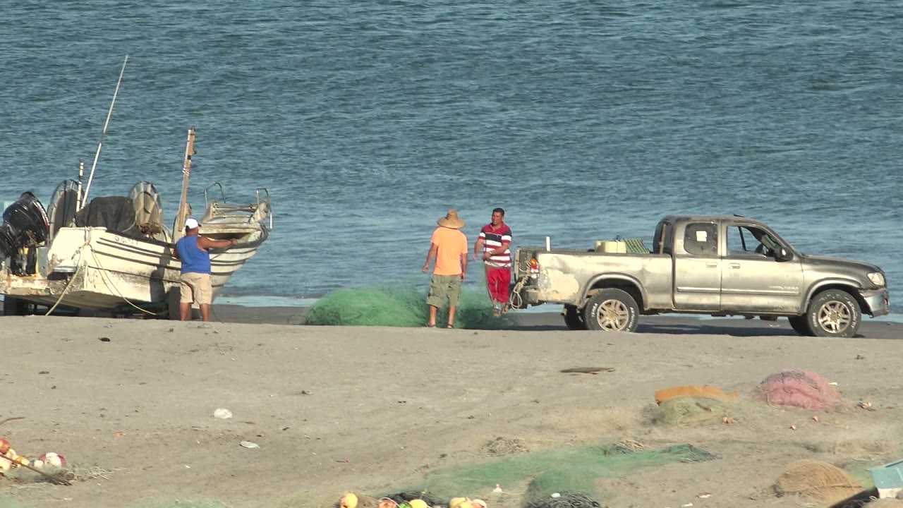 Men stand beside a pickup truck and green nets on the beach near a boat.
