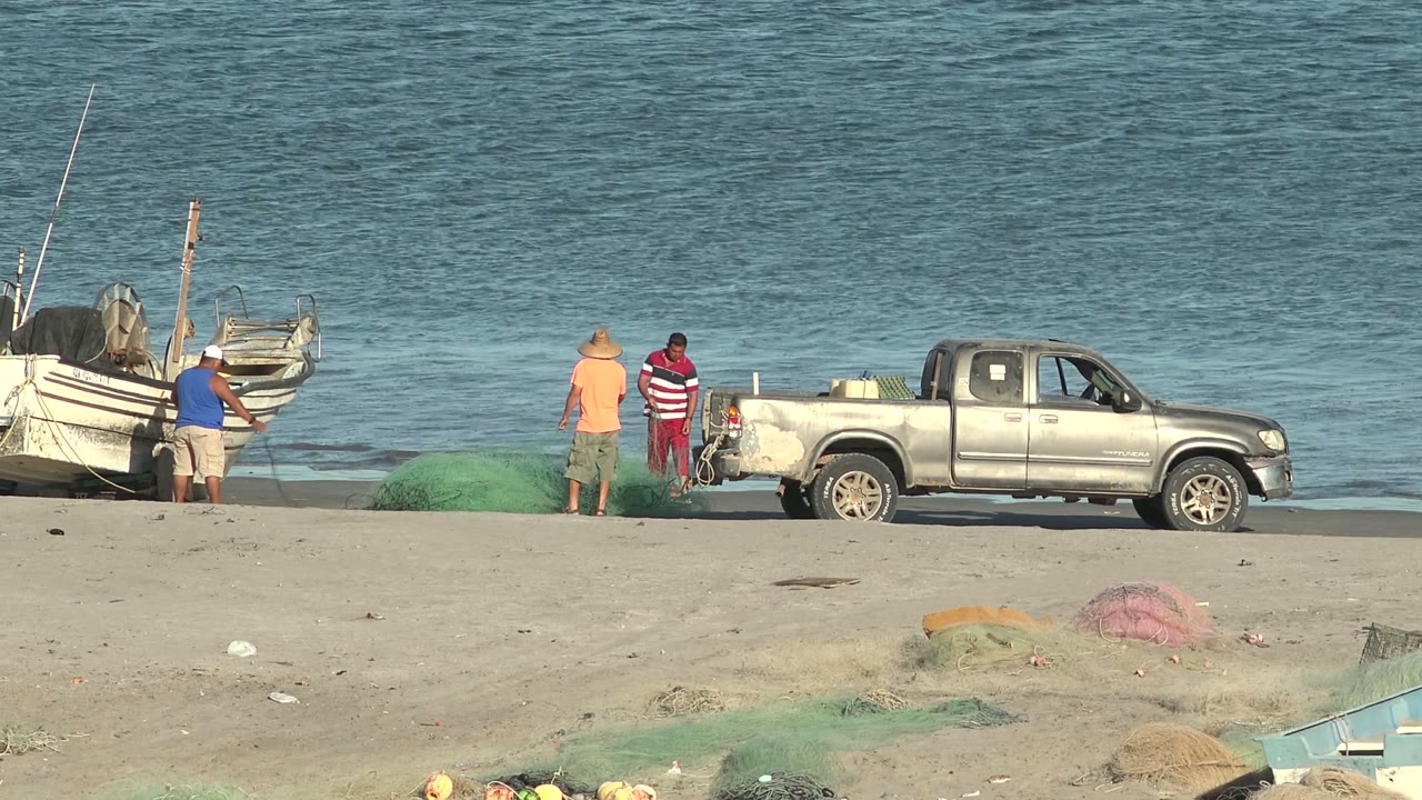 At 0:02, two men stand near the truck bed and one remains by the boat while green nets lie across the sand in San Juanico, Baja California Sur.