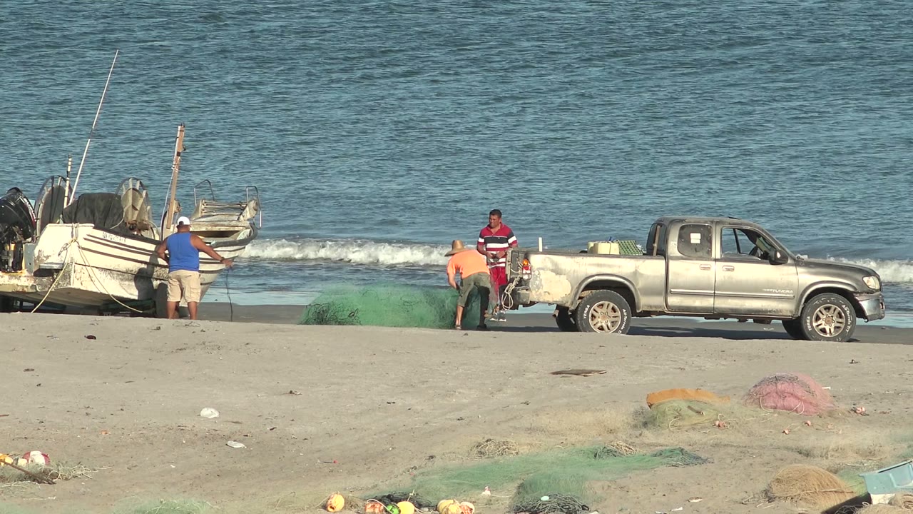 Fishermen work near green nets, a silver pickup, and small white boats on a sandy beach in San Juanico, Baja California Sur, with blue ocean water behind them.