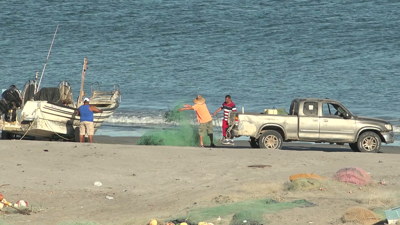 Man in orange shirt lifts green fishing net near a pickup truck and boats.