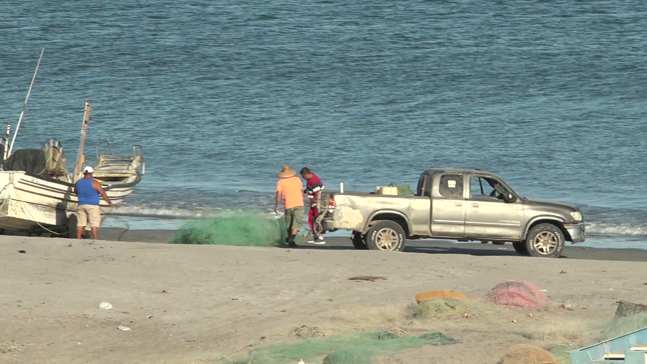 Several fishermen stand near the rear of a silver pickup and green netting on the sand, with small boats at the shoreline and ocean water behind them in San Juanico.