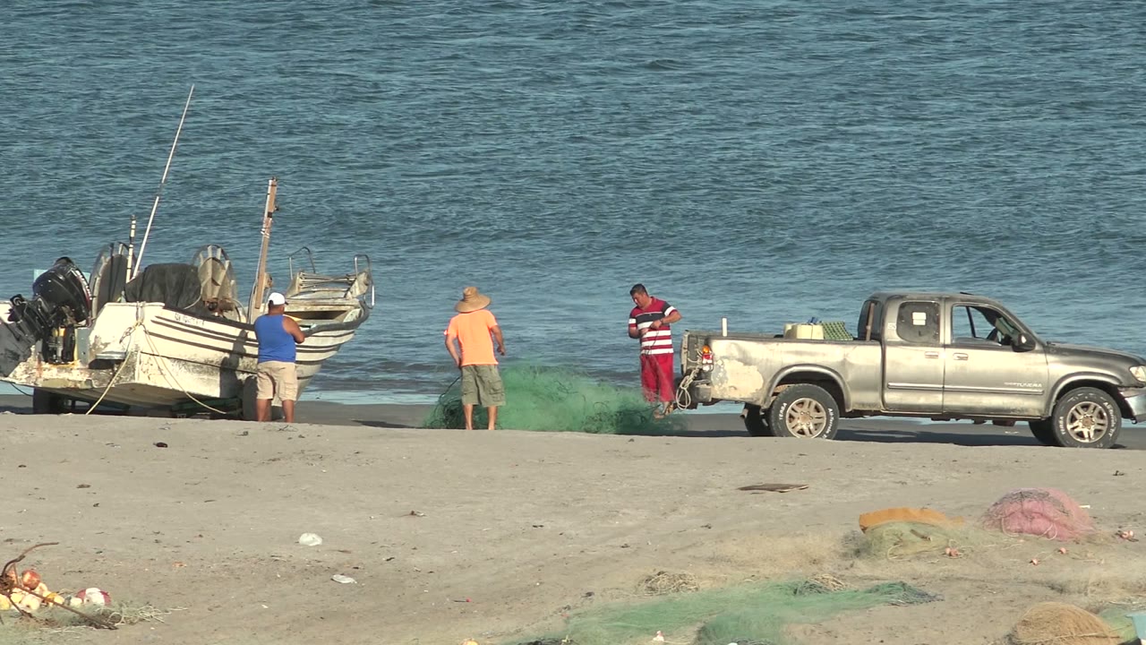 At 0:00 in San Juanico, Baja California Sur, three men stand near green fishing nets, a silver pickup truck, and small boats on a sandy beach with blue ocean behind them.