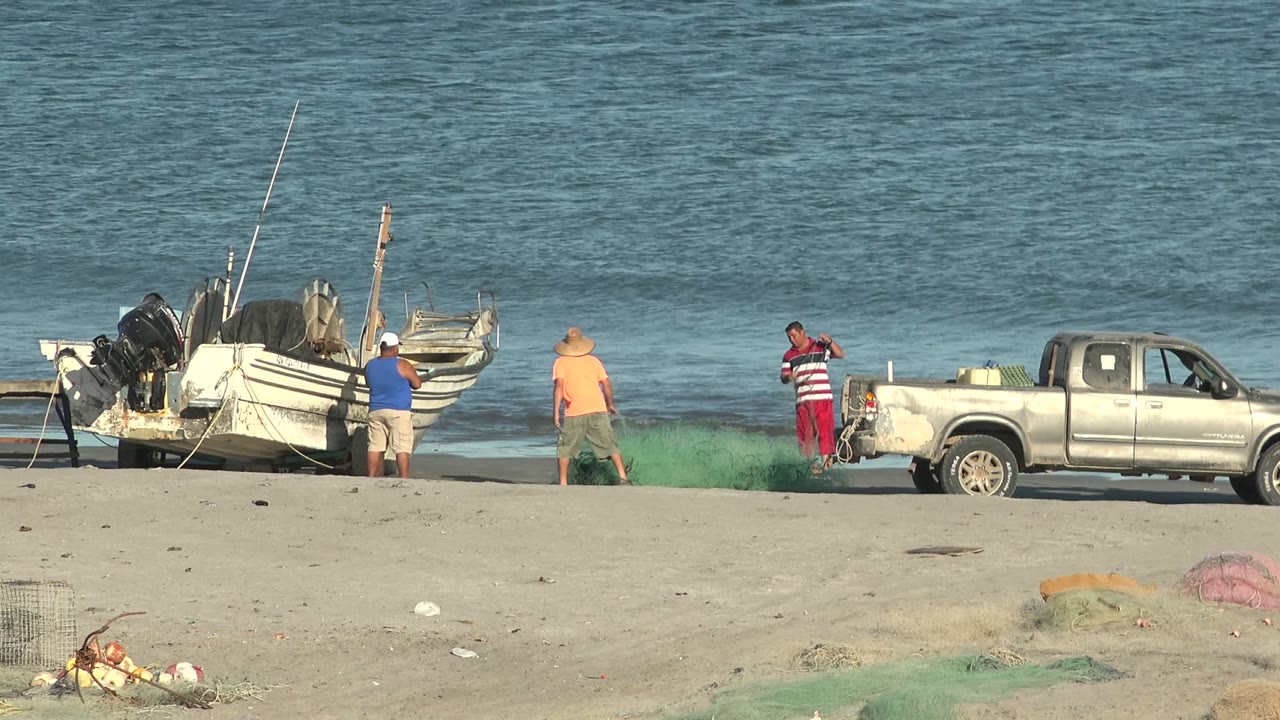 Men adjust fishing nets beside a pickup truck and boats at the shoreline.