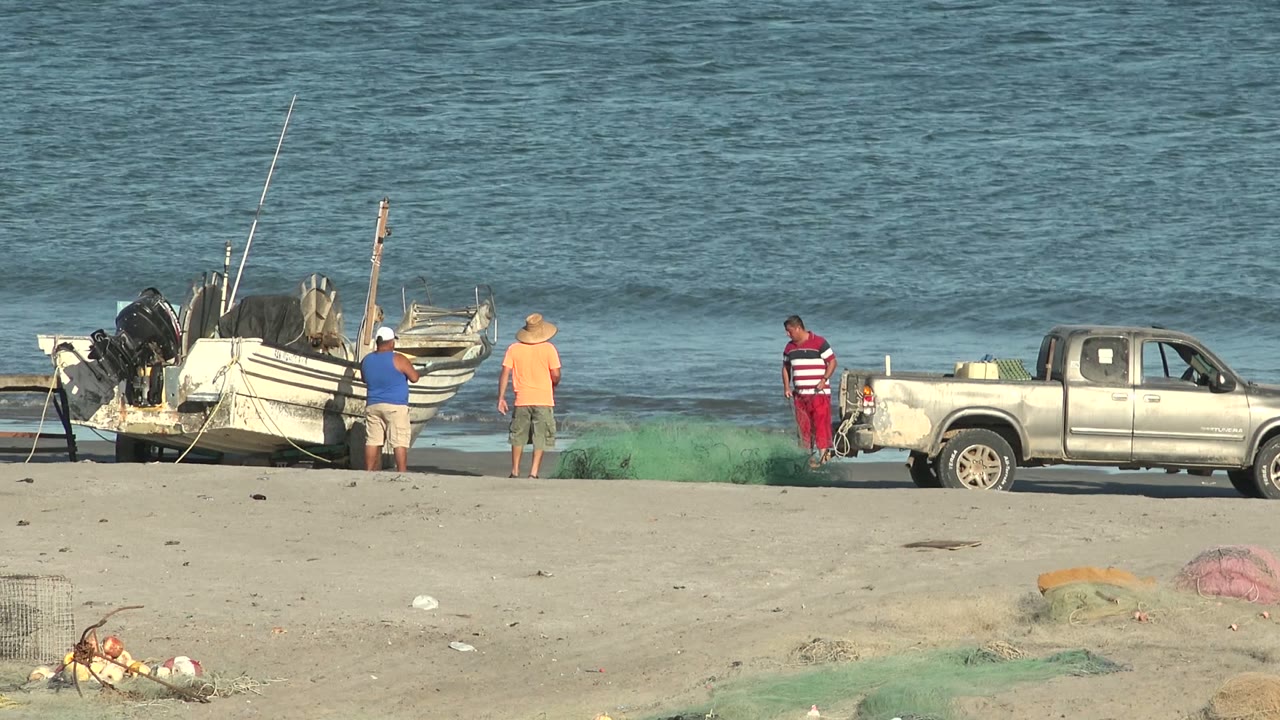 At 0:02 in San Juanico, Baja California Sur, the men remain near the truck and nets while the boats sit at the left edge and a small wave line forms behind them.
