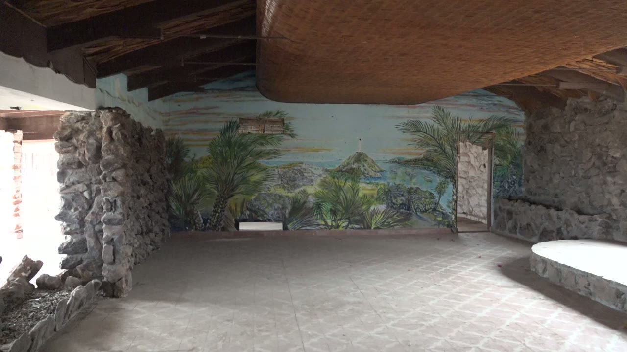 Wide view inside a stone-walled room in Mulege, Baja California Sur, with a tropical mural on the back wall, woven ceiling overhead, pale concrete floor, and no visible people.