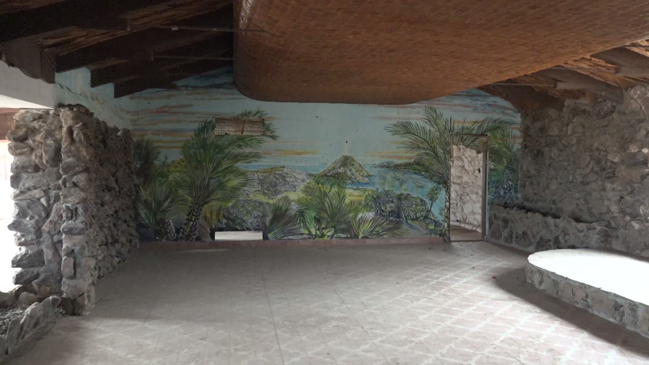 The pan continues inside the Mulege room, with the tropical mural spanning the back wall, a narrow doorway at right, a curved stone bench edge in the foreground, and no visible people.