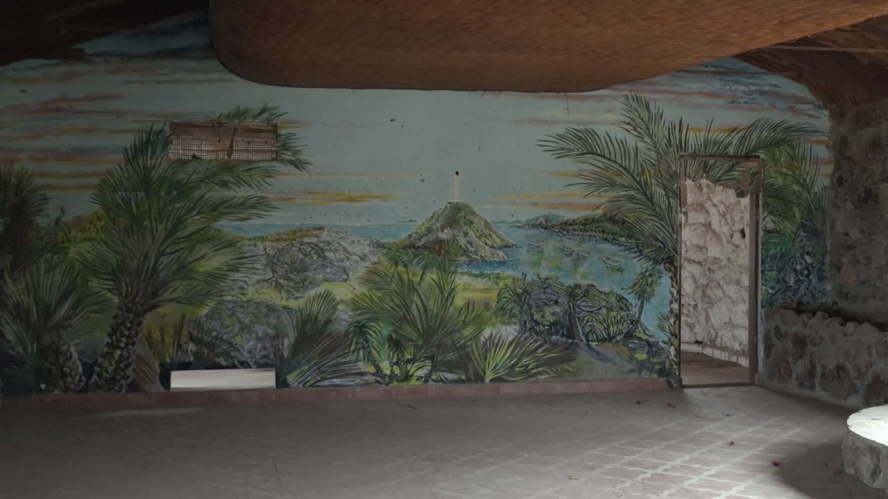 Wide view inside a Mulege, Baja California Sur room with a tropical island mural across the wall, a small white sink at lower left, tiled floor, dark ceiling beams, and no visible people.