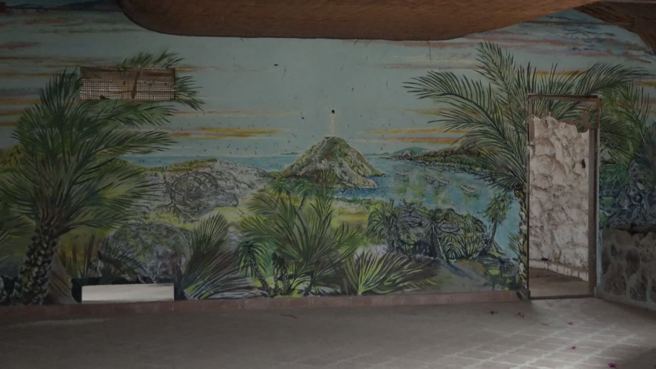 The mural fills more of the Mulege room frame, with a painted island and shoreline centered between palms, a stone doorway at right, dark ceiling beams above, and no visible people.