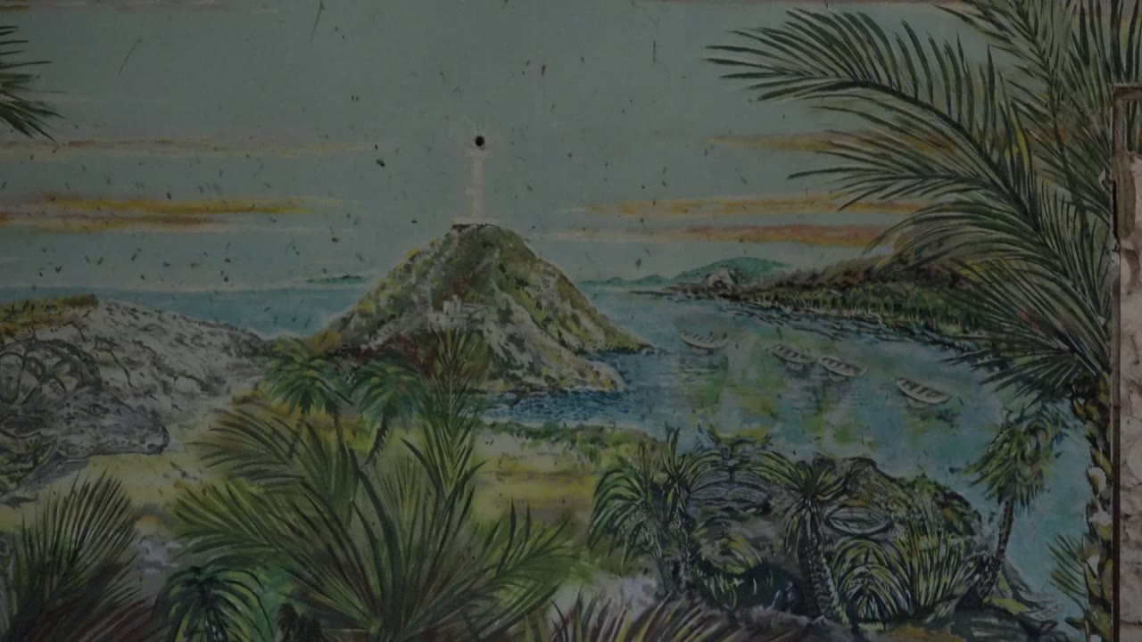 At 0:02 in Mulege, Baja California Sur, the lighthouse beacon area is visible above the rocky island while palm fronds arc across the right side, with boats on the water and no visible people.