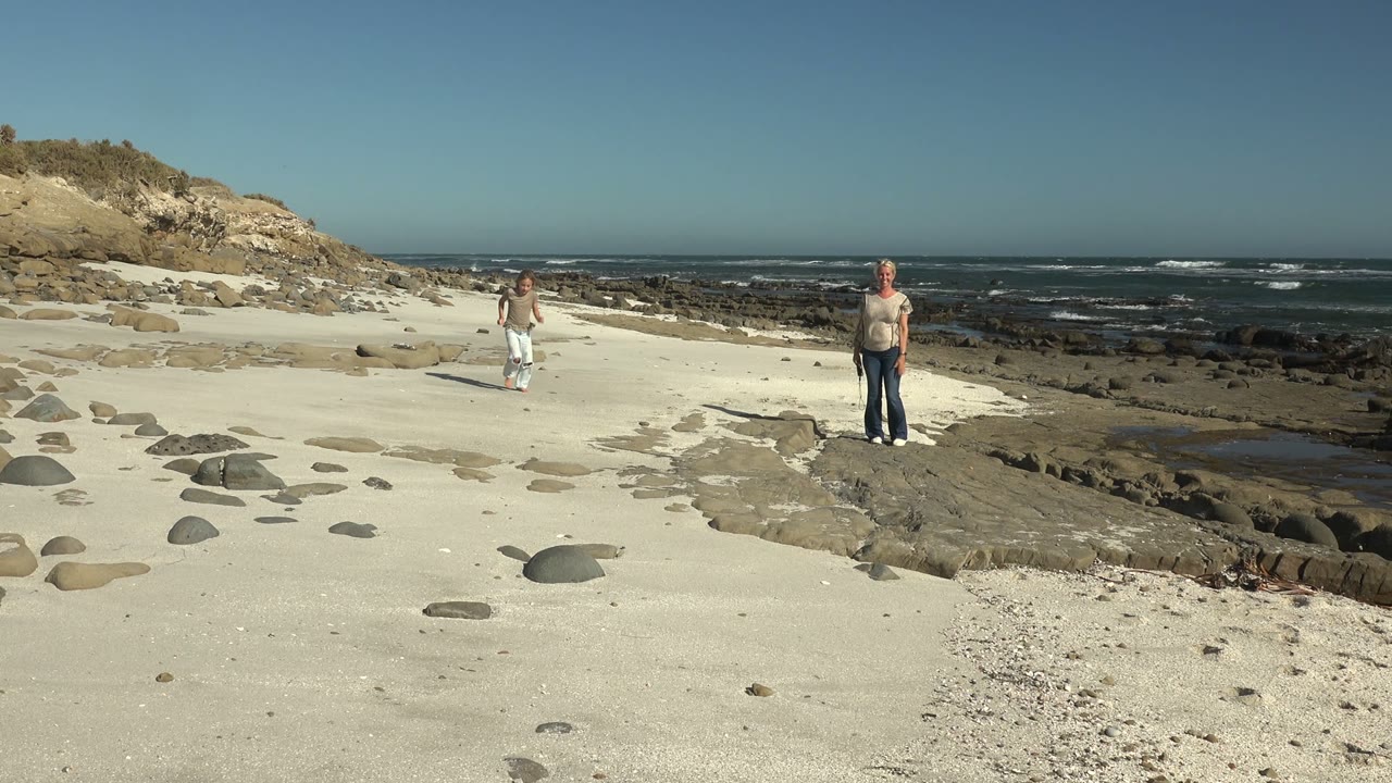 At 0:00 in La Bocana, Mexico, a child and adult are separated across the sandy beach while rocks, surf, and a low bluff fill the background.
